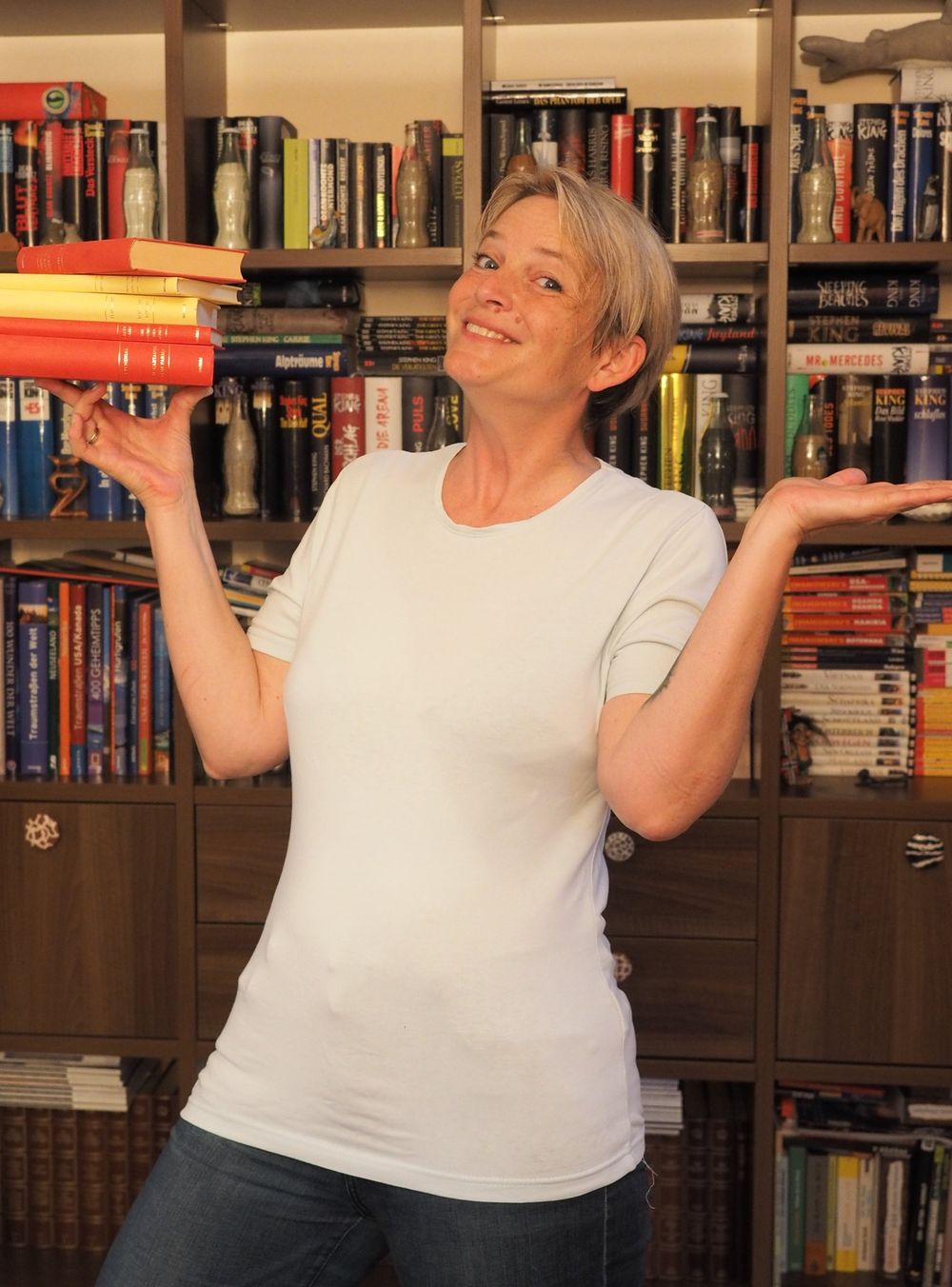 Andrea Wiesauer lächelt vor einem Bücherregal und balanciert einen Bücherstapel auf der Hand
